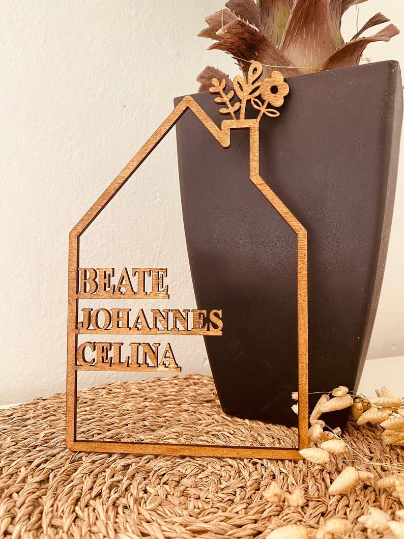 May include: Wooden house-shaped sign with the names "Beate", "Johannes", and "Celina" inscribed on it. The sign has a small flower design at the top.