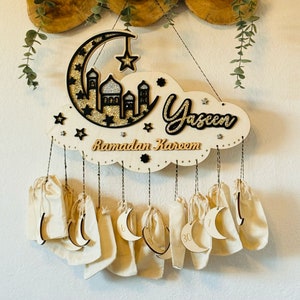 May include: A wooden Ramadan advent calendar with a crescent moon and mosque design. The calendar has 30 small pouches hanging from it, each with a number printed on it. The calendar is decorated with gold glitter and the words "Ramadan Kareem" and "Yaseen".