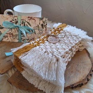 Puede incluir: Un juego de posavasos de macramé hechos a mano en blanco y amarillo mostaza, con detalles de flecos. Una tarjeta floral atada con una cinta azul y una taza de té están en el fondo. Una pequeña etiqueta dice "Handmade with love".