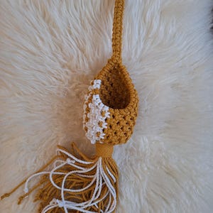 May include: Mustard yellow macrame plant hanger with a white and yellow woven design. The plant holder has a wooden ring at the top for hanging and a tassel at the bottom. The macrame is made of thick, soft yarn.