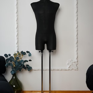 Può includere: Un busto di manichino nero dal design elegante e minimalista. La forma poggia su due pali metallici color argento fissati a una base rettangolare nera. Il manichino è posto contro un muro bianco con modanature decorative.