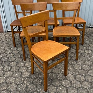 Könnte beinhalten: Sieben Holzstühle mit hellbraunem Finish. Jeder Stuhl hat eine rechteckige Sitzfläche, eine Sprossenlehne und vier Beine. Die Stühle sind in einer Gruppe angeordnet und zeigen ihr schlichtes, klassisches Design.