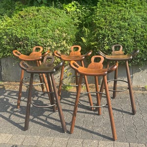Può includere: Un gruppo di cinque sgabelli da bar in legno con sedili curvi e una piccola maniglia nella parte superiore dello schienale. Gli sgabelli hanno gambe marrone scuro e un poggiapiedi in metallo. Gli sgabelli sono disposti su una superficie pavimentata con fogliame verde sullo sfondo.