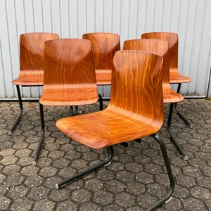 Op de afbeelding: Een set van zes houten stoelen met een warme, roodbruine afwerking. De stoelen hebben gebogen rugleuningen en zittingen en worden ondersteund door donkergrijze metalen poten. De stoelen staan op een getextureerd, grijs oppervlak.