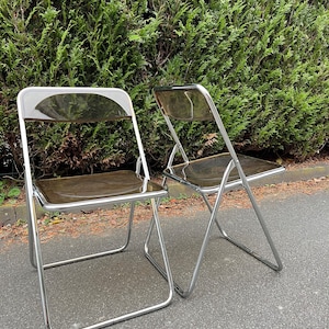 Könnte beinhalten: Zwei Klappstühle mit transparenten Sitzen und Rückenlehnen und verchromten Metallrahmen. Die Stühle stehen auf einer gepflasterten Fläche vor einer dichten grünen Hecke. Modernes Design.