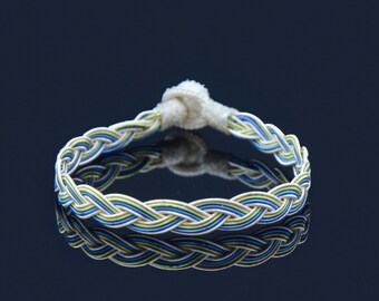 May include: A braided bracelet with blue, green, and white cords. The bracelet is tied with a white knot.
