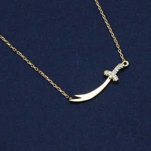 May include: A gold chain necklace with a small, curved sword pendant encrusted with clear cubic zirconia stones.