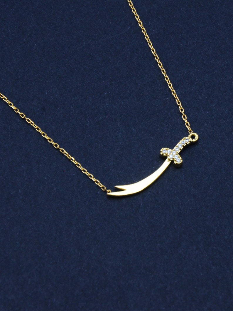May include: A gold chain necklace with a small, curved sword pendant encrusted with clear cubic zirconia stones.