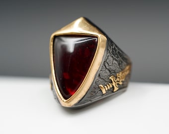 May include: A striking ring featuring a large, triangular, deep red gemstone set in a gold-toned bezel. The ring's band is textured, with a dark finish and gold-toned decorative elements. A unique piece of jewelry.