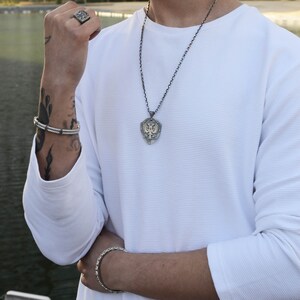 May include: A silver chain necklace with a shield-shaped pendant featuring a detailed eagle design. The pendant has a gold-toned finish. The necklace is worn by a person with a white long-sleeved shirt.