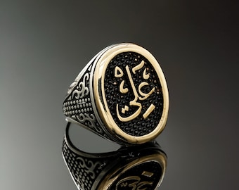 May include: A silver ring with a gold-colored oval centerpiece. The centerpiece features Arabic calligraphy on a black textured background. The ring has a detailed, patterned band and is reflected on a dark surface.