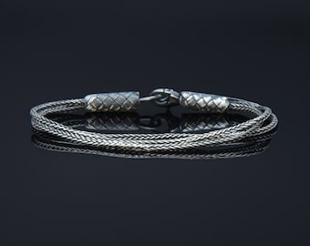 May include: A silver bracelet with a braided chain design and woven end caps. The bracelet is on a black background.