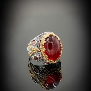 Könnte beinhalten: Ein silberner Ring mit einem großen roten Edelstein, umgeben von goldenen Akzenten und kleineren roten Edelsteinen.