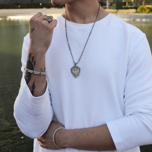 May include: A silver chain necklace with a shield-shaped pendant featuring a detailed design. The pendant is worn by a person with a white long-sleeved shirt and a silver bracelet on their wrist.