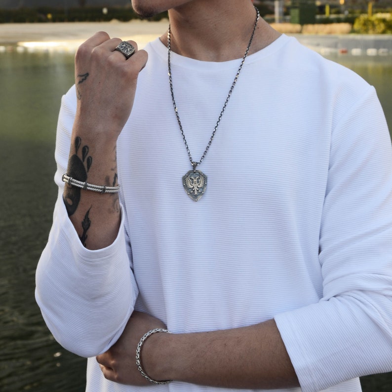 May include: A silver chain necklace with a shield-shaped pendant featuring a detailed design. The pendant is worn by a person with a white long-sleeved shirt and a silver bracelet on their wrist.
