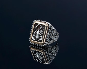 May include: A silver ring with a gold-colored rectangular face featuring a scorpion design. The ring has a detailed pattern on the band and is set against a dark background, creating a reflective surface.