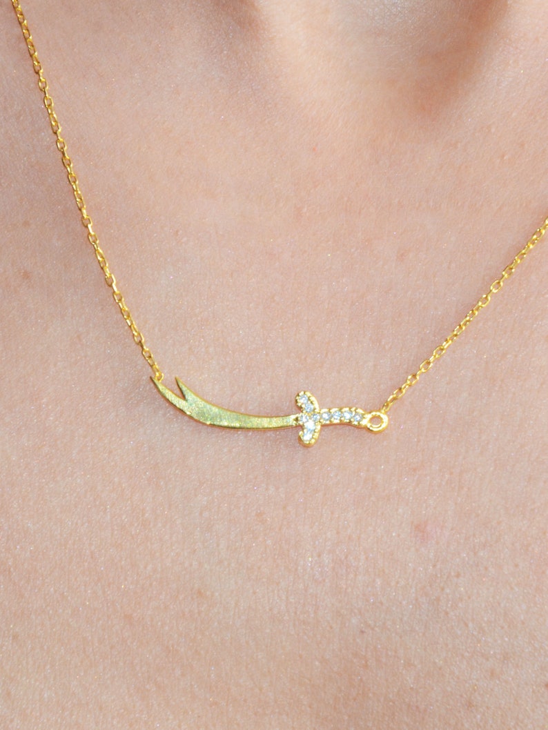 May include: A gold necklace with a small, diamond-encrusted sword pendant.