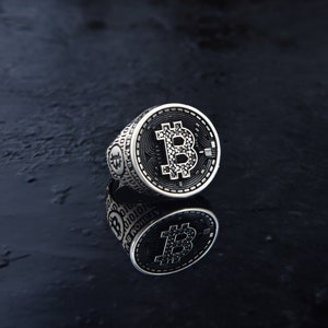 Puede incluir: Un anillo de plata con un diseño de bitcoin negro y plateado. El anillo está sobre una superficie negra y se refleja en la superficie.