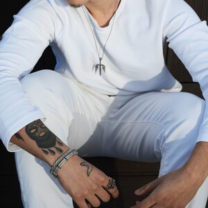 May include: A person wearing a white long-sleeved shirt with a silver necklace featuring a winged sword pendant. The person is wearing a silver bracelet on their left wrist and a silver ring on their right hand. The person has a black and white tattoo on their left arm.
