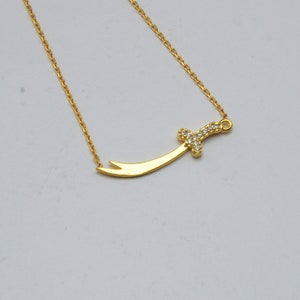 May include: A gold chain necklace with a small sword pendant encrusted with clear cubic zirconia stones.