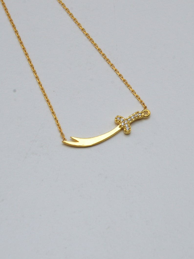 May include: A gold chain necklace with a small sword pendant encrusted with clear cubic zirconia stones.
