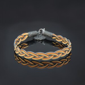 Puede incluir: Un brazalete trenzado con bandas naranja, amarilla y gris. El brazalete tiene un cierre plateado.