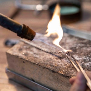 May include: A close-up of a silver chain being heated with a torch. The chain is held in place with tweezers on a rough, gray surface.