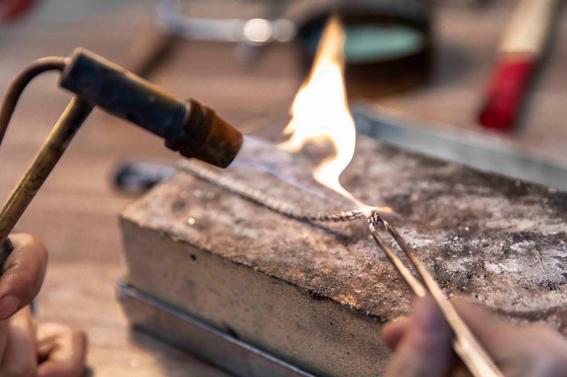 May include: A close-up of a silver chain being heated with a torch. The chain is held in place with tweezers on a rough, gray surface.