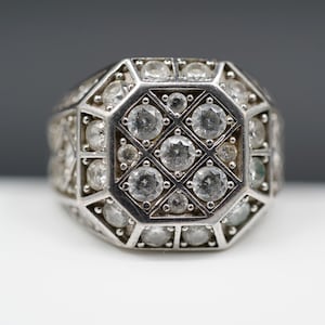 Könnte beinhalten: Ein silberfarbener Ring mit geometrischem Design, besetzt mit zahlreichen klaren, runden Edelsteinen. Die Ringoberfläche hat ein zentrales quadratisches Muster, umgeben von achteckigen Formen, alle besetzt mit den funkelnden Steinen.
