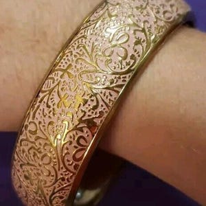 May include: A pink and gold bangle bracelet with an ornate floral design. The bracelet has a gold-colored metal base and a pink enamel overlay with a detailed, repeating pattern. The design features swirling lines and floral motifs.