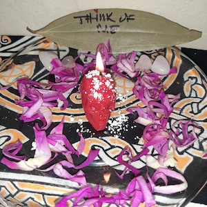 May include: A red candle with white powder on top, surrounded by pink flower petals on a black and orange patterned surface. The text "Think of me" is written on a green leaf.