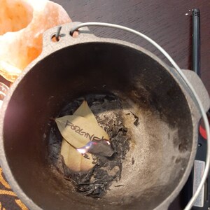 May include: A black cast iron pot with a handle, containing a bay leaf with the word "Forgive" written on it. The leaf is partially burned and there is ash in the bottom of the pot.