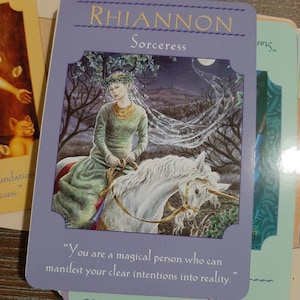 May include: A purple card with the text "Rhiannon Sorceress" and an illustration of a woman riding a white unicorn through a forest. The text below the image reads "You are a magical person who can manifest your clear intentions into reality."