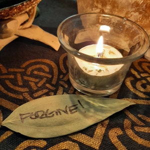May include: A lit candle in a glass jar sits on a dark brown surface with a leaf that says "Forgive!" written on it.