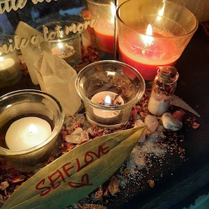 May include: A close-up of a spell jar with a bay leaf with the word "Self Love" written on it. The jar is surrounded by candles, crystals, and rose petals.