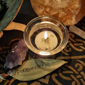 May include: A glass candle holder with a white candle inside. The candle has dried herbs and flowers embedded in it. The candle is lit and the flame is visible. The candle holder is sitting on a dark surface with a green leaf and a purple crystal.