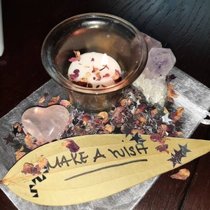 May include: A pink heart-shaped crystal, a small glass candle holder with a white candle, and a purple crystal are arranged on a silver and white background with a leaf-shaped tag that says "Make a Wish".