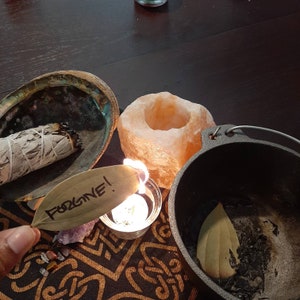 May include: A close-up of a salt lamp with a small candle burning inside. A bay leaf is in a black cast iron pot. A piece of sage is on a wooden surface. A leaf with the word "Forgive!" written on it is in the foreground.