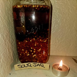 Op de afbeelding: Een glazen pot gevuld met een donkere vloeistof en rode pepervlokken, met het label "SOUR JAR". Een kleine brandende kaars staat naast de pot. De pot lijkt een weckpot te zijn.
