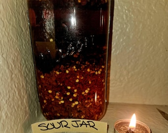 May include: A glass jar containing a dark liquid and red pepper flakes, with a label reading "SOUR JAR." A small lit candle is positioned beside the jar. The jar appears to be a mason jar.