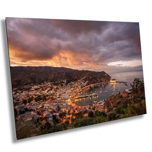 May include: A high-angle view of a coastal town nestled in a bay at twilight. The scene features illuminated buildings, a harbor filled with small boats, and a large circular building on the water, all set against a dramatic orange and purple sunset.