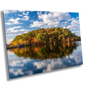 May include: A scenic view of a lake with a small island covered in colorful autumn foliage. The trees are a mix of red, orange, yellow, and green. The sky is blue with white clouds reflected in the still water.