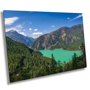 May include: A landscape print showcasing a turquoise lake nestled amidst dense green forests and majestic mountains under a clear blue sky with fluffy clouds. The image depicts a tranquil natural scene.