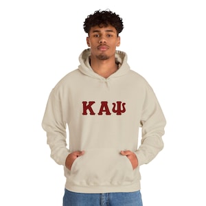 May include: A sand-coloured hooded sweatshirt featuring the Greek letters Kappa Alpha Psi in a dark red, collegiate-style font across the chest. The garment includes a front kangaroo pocket, long sleeves with ribbed cuffs, and a matching drawstring hood.