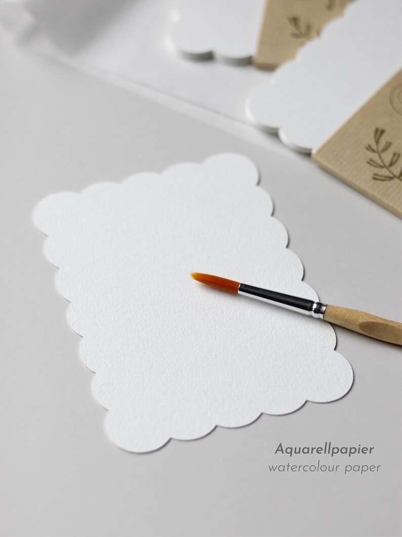 Postkarte aus Aquarellpapier mit stark gewelltem Rand liegt neben einem Stapel an fertig verpackten Postkarten-Sets