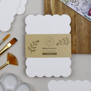 Postkartenset aus Aquarellpapier mit stark gewelltem Rand liegt neben einem Stapel an fertig verpackten Postkarten-Sets, verschiedenen Pinseln und einer Farbpalette