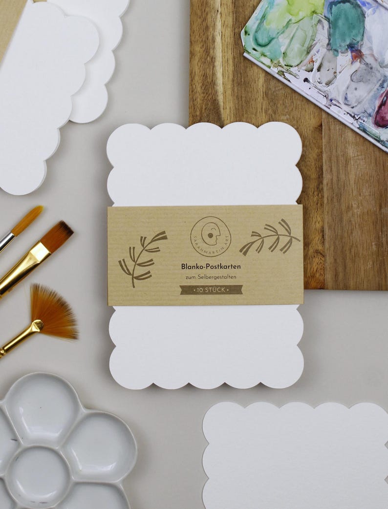 Postkartenset aus Aquarellpapier mit stark gewelltem Rand liegt neben einem Stapel an fertig verpackten Postkarten-Sets, verschiedenen Pinseln und einer Farbpalette