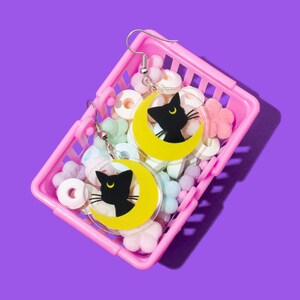 Puede incluir: Un par de pendientes de gato negro y luna creciente en una cesta de la compra de pl&aacute;stico rosa llena de caramelos de colores pastel.