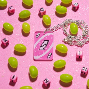 May include: A pink and white card with a reversed arrow symbol on it. The card is hanging from a silver chain necklace. The card is surrounded by green jelly beans on a pink background.