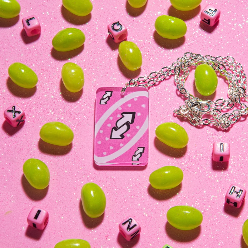 May include: A pink and white card with a reversed arrow symbol on it. The card is hanging from a silver chain necklace. The card is surrounded by green jelly beans on a pink background.